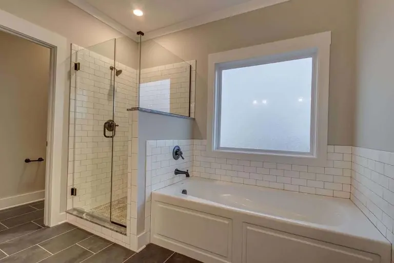 A white bathtub in front of a picture window adjoined by a custom tile shower with a glass door.