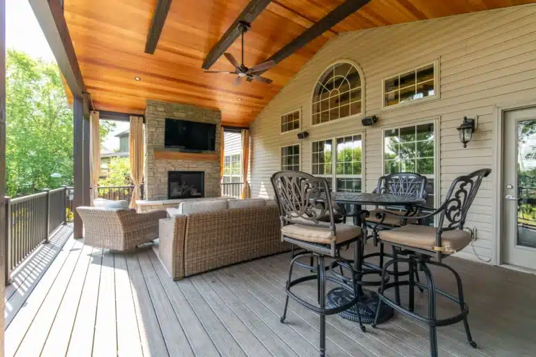 Covered deck in Lexington, KY with a stone fireplace and mounted TV, wicker sofa and chairs, and a wrought-iron dining set with bar stools.