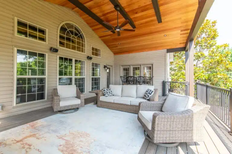 Covered outdoor deck in Lexington, KY with a cedar plank ceiling, ceiling fan, wicker sofa and chairs on an area rug, overlooking a wooded backyard.