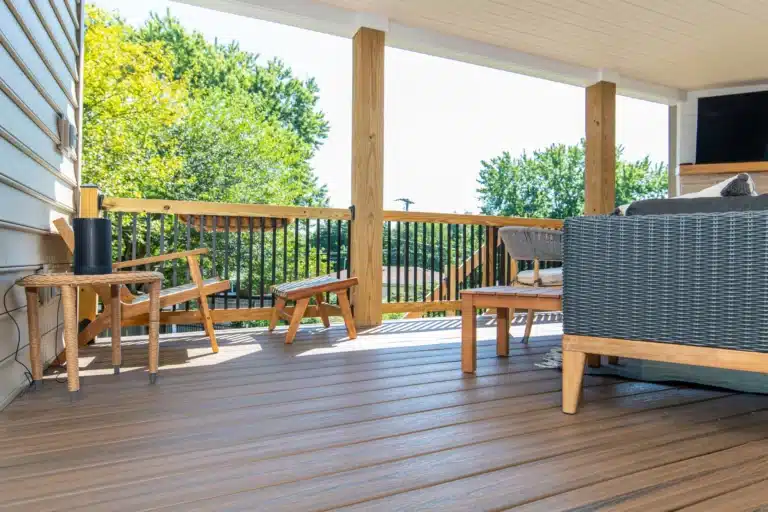 Covered deck corner with wooden side tables, a bench, and wicker seating, overlooking a tree-lined yard.