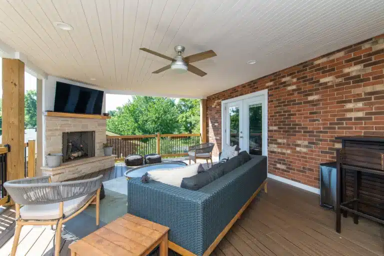 Covered deck with a brick wall, stone fireplace, mounted TV, and a dark wicker sofa with cushions.