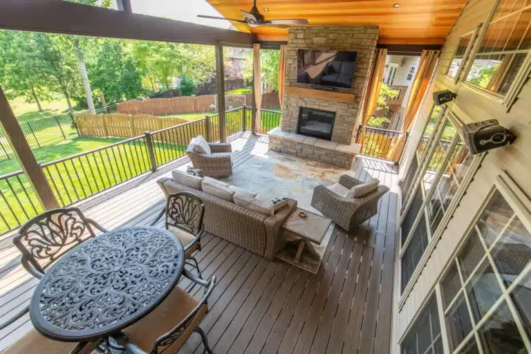 Covered deck seating area with a stone fireplace, wall-mounted TV, wicker furniture, and a view of the fenced backyard.