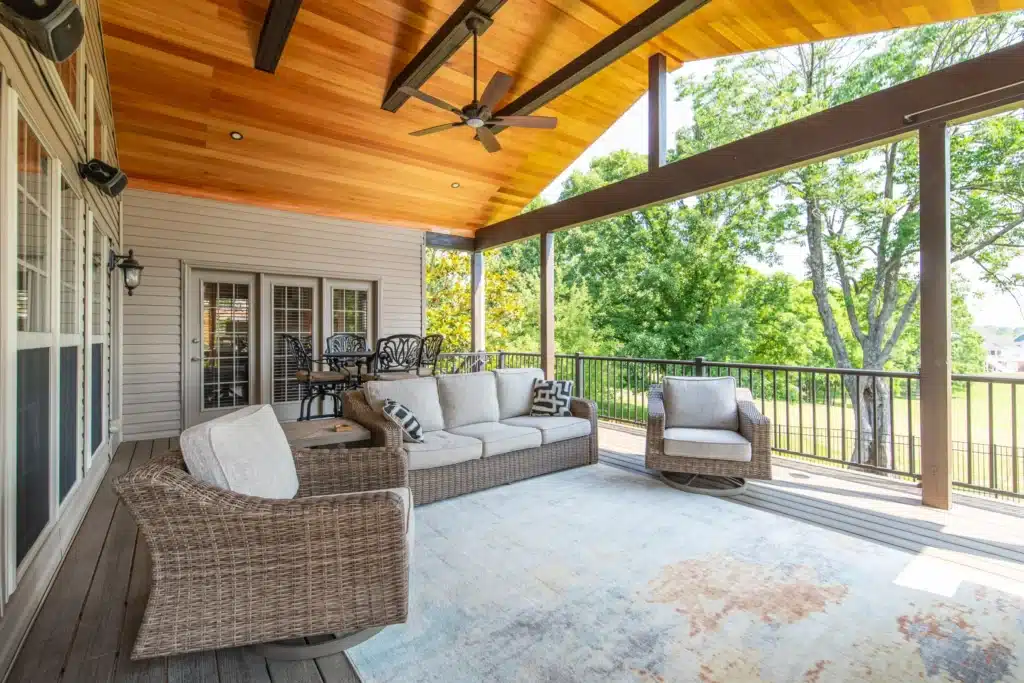 Covered deck lounge area with wicker sofa and chairs on a rug, overlooking a grassy yard lined with trees.
