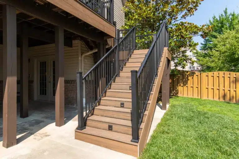 Composite wood staircase with black metal railing leading up to a deck, next to a covered patio and backyard.